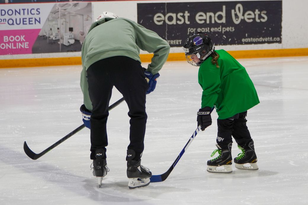 A camper in green being shown how to hold a stick while standing still on the ice