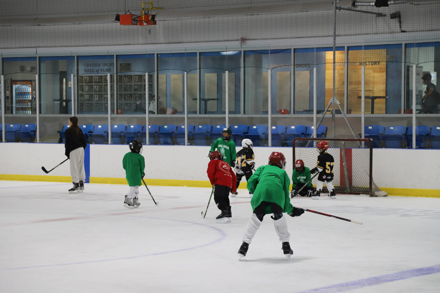 A game in progress at Leaside Arena, campers in green and red jerseys pressing the net with the arena stand and the Leaside Sports Hall of Fame wall behind them
