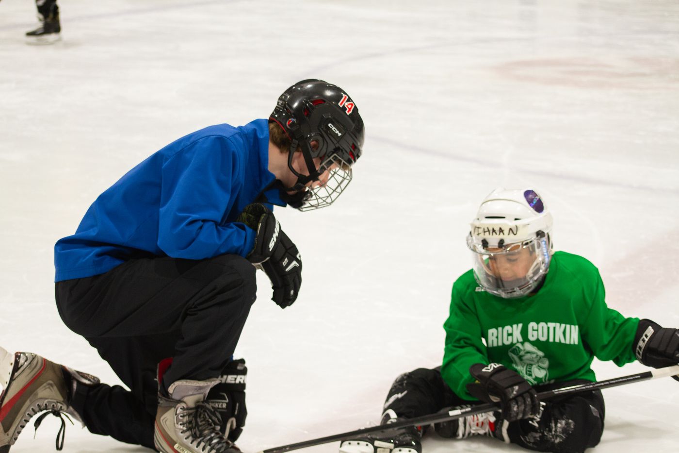 A volunteer in a blue jacket crouching on the ice to help a fallen camper back onto their skates