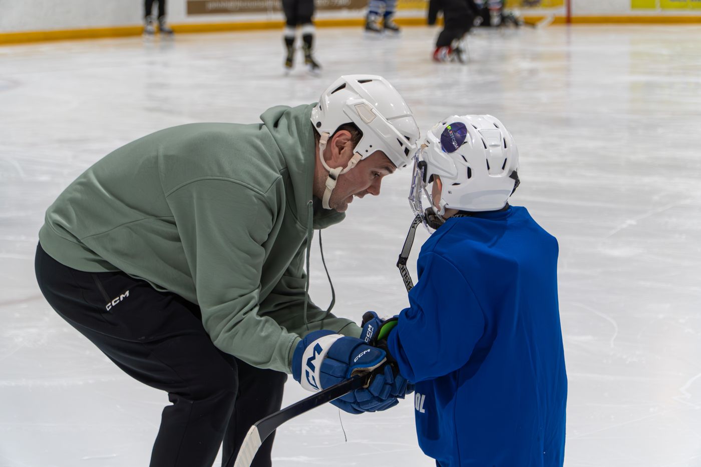 A coach in a green hoodie bent down to demonstrate a stick grip to a young camper in blue