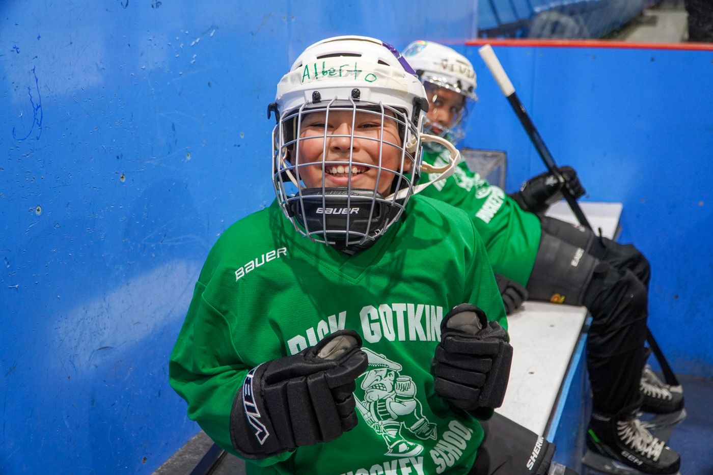 A camper in a green jersey and white helmet smiling on the ice while holding a stick